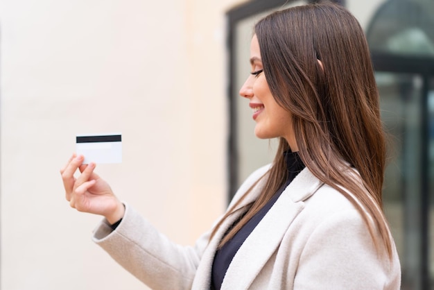 Mujer bonita joven sosteniendo una tarjeta de crédito al aire libre con expresión feliz