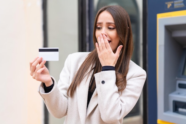 Mujer bonita joven sosteniendo una tarjeta de crédito al aire libre con sorpresa y expresión facial conmocionada