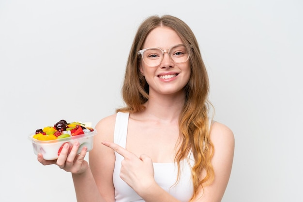 Mujer bonita joven sosteniendo un tazón de fruta aislado sobre fondo blanco y apuntándolo