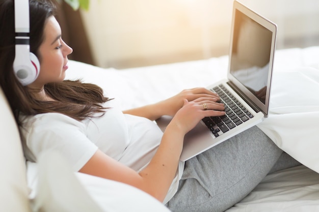 Mujer bonita joven tiene un descanso en la cama con una computadora portátil y una lista de música