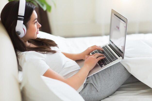 Mujer bonita joven tiene un descanso en la cama con una computadora portátil y una lista de música