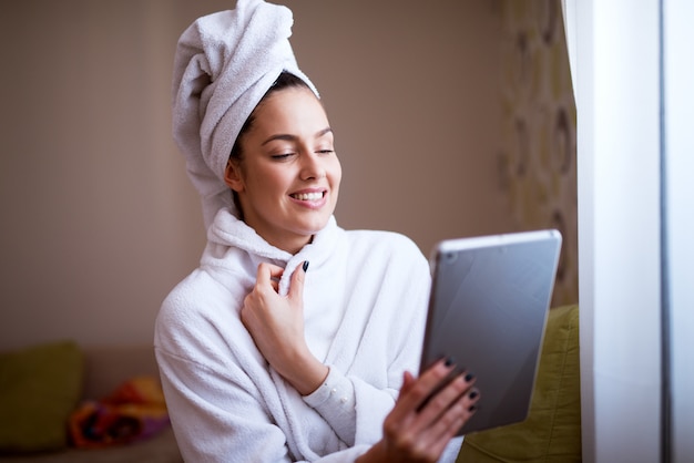 La mujer bonita joven en traje está sonriendo y sintiéndose fresca después de la ducha mientras que usa la tableta cerca de una ventana.