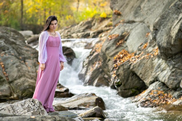 Mujer bonita joven en vestido largo de moda de pie cerca de un pequeño río de montaña con agua en movimiento rápido.