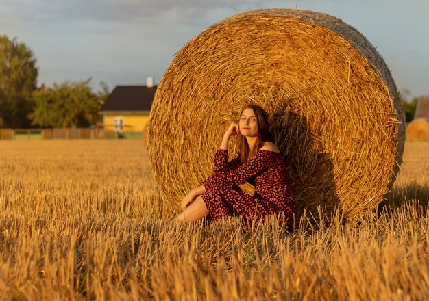 Mujer bonita joven en vestido rojo de cerca en el fondo del campo con un pajar al atardecer.