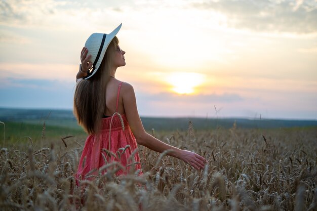 Mujer bonita joven en vestido rojo de verano y sombrero de paja caminando en el campo de cultivo amarillo con trigo dorado maduro disfrutando de la cálida noche.