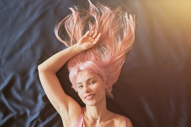 Mujer bonita con largo cabello rosado suelto y joyas de plata se encuentra en una cama suave y cómoda