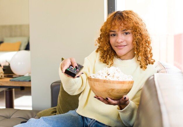 Mujer bonita latina de pelo rojo joven comiendo palomitas de maíz en un sofá y viendo una película