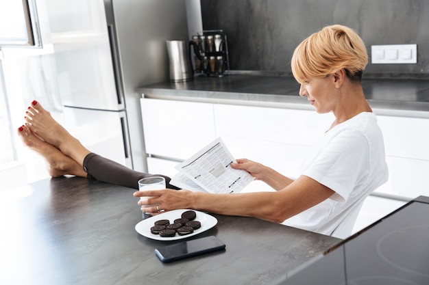 Mujer bonita leyendo un periódico sentado en la mesa de la cocina, bebiendo leche de un vaso, comiendo galletas