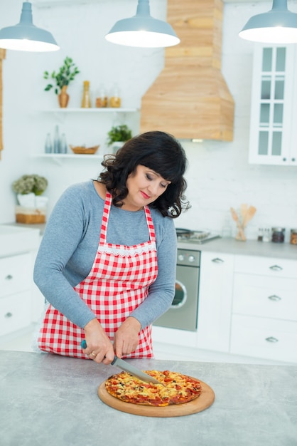 Mujer bonita madura corta pizza readymade con un cuchillo en la cocina Cocinar y quehaceres domésticos