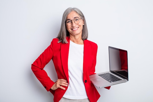 Mujer bonita mayor que sonríe feliz con una mano en la cadera y confiada. concepto de negocio y portátil