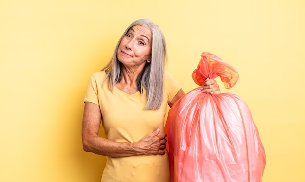 Mujer bonita de mediana edad encogiéndose de hombros, sintiéndose confusa e insegura. bolsa de plástico para garbaje