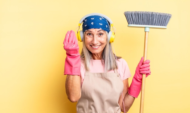 Mujer bonita de mediana edad haciendo capice o gesto de dinero, diciéndole que pague. concepto de hogar y escoba