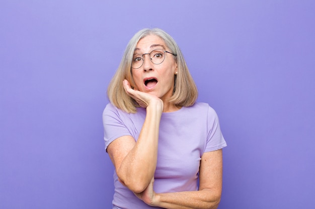 Mujer bonita de mediana edad o mayor con la boca abierta en estado de shock e incredulidad, con la mano en la mejilla y el brazo cruzado, sintiéndose estupefacta y asombrada