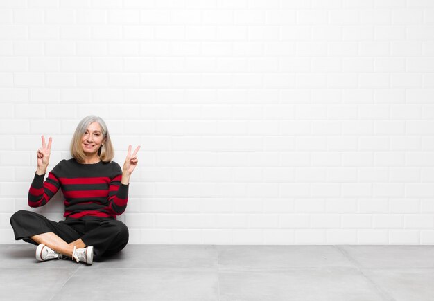 Mujer bonita de mediana edad o mayor sonriendo y mirando feliz, amigable y satisfecha, gesticulando victoria o paz con ambas manos