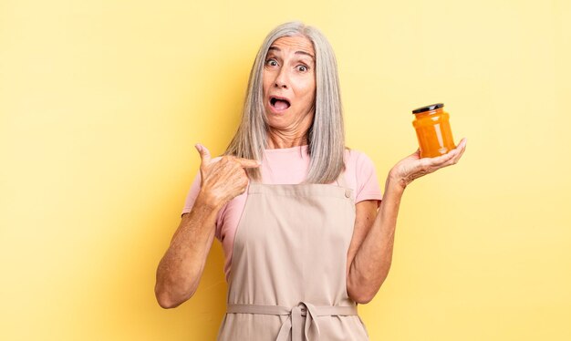 Mujer bonita de mediana edad que parece sorprendida y sorprendida con la boca abierta apuntando a la mermelada de melocotón