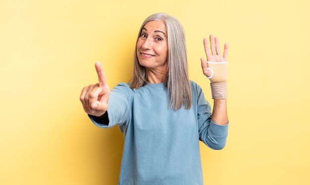 Mujer bonita de mediana edad sonriendo con orgullo y confianza haciendo el número uno. concepto de vendaje de mano