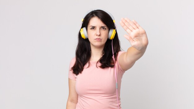 Mujer bonita mirando seria mostrando la palma abierta haciendo gesto de parada escuchando música con auriculares