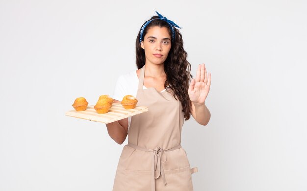 Mujer bonita mirando seria mostrando la palma abierta haciendo gesto de parada y sosteniendo una bandeja de muffins
