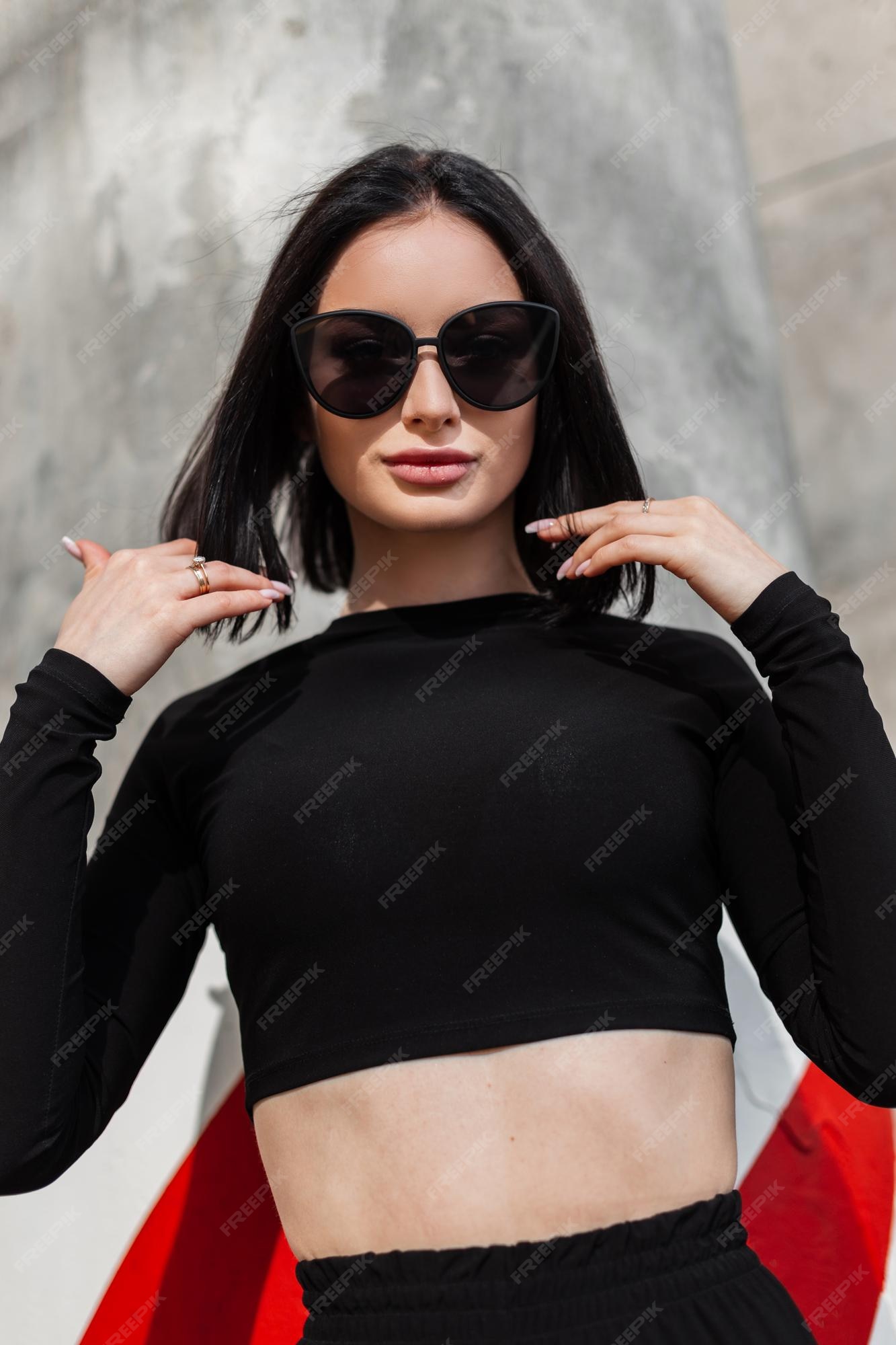 Mujer bonita de moda con elegantes gafas de sol en traje deportivo negro con top de manga larga camina cerca de muro de hormigón la calle | Foto Premium