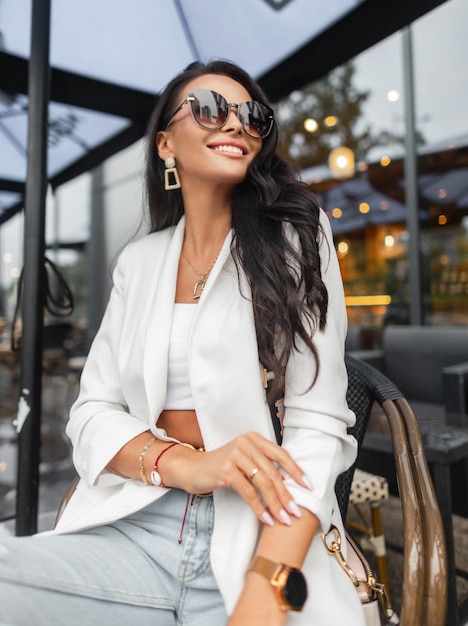 Una mujer bonita a la moda con una sonrisa en un atuendo informal de negocios de moda con un blazer blanco superior y jeans se sienta y disfruta en una silla en un café en la calle