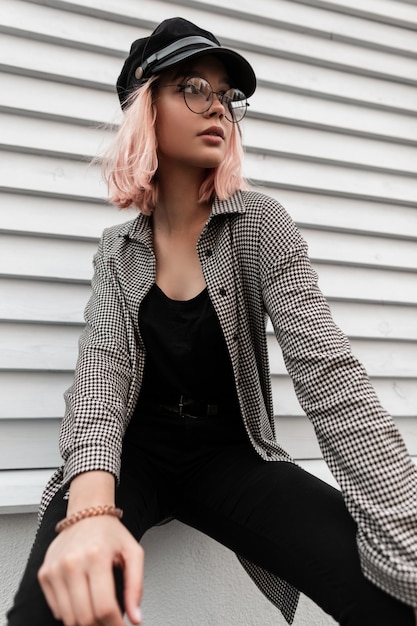 Mujer bonita modelo hipster con gafas elegantes y una gorra de moda en ropa de mezclilla con una camisa a cuadros se sienta cerca de una pared de madera