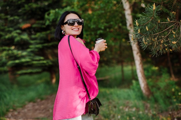 Mujer bonita morena con gafas de sol sosteniendo una taza de café y caminando en el parque