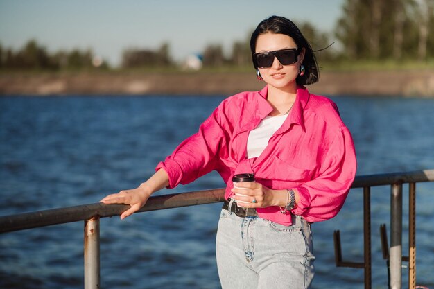 Mujer bonita morena con gafas de sol con taza de café en el muelle al borde del lago