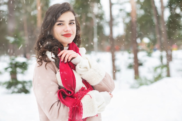 Mujer bonita morena lleva abrigo y bufanda roja caminando en el bosque posando durante las nevadas. Espacio para texto