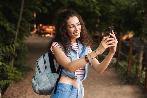 Mujer bonita morena con mochila, sonriendo y fotografiando la naturaleza en el teléfono inteligente mientras camina por el parque verde