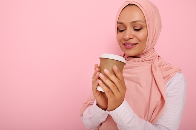 Mujer bonita musulmana árabe con la cabeza cubierta en hijab rosa bebiendo bebida caliente, té o café de una taza de cartón desechable para llevar, de pie tres cuartos contra el fondo de color con espacio de copia