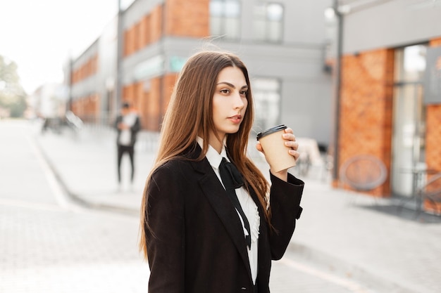 Mujer bonita de negocios con traje negro de moda camina por la ciudad y bebe café