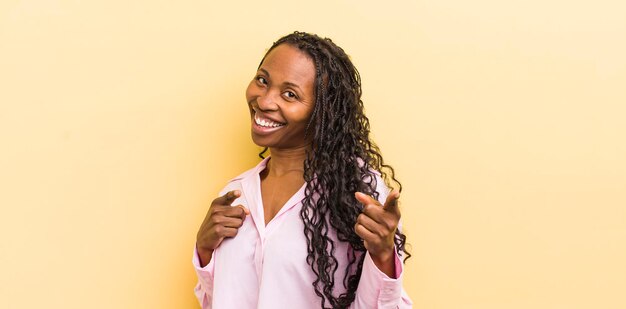 Mujer bonita negra sonriendo con una actitud feliz exitosa positiva apuntando a la cámara haciendo un signo de pistola con las manos