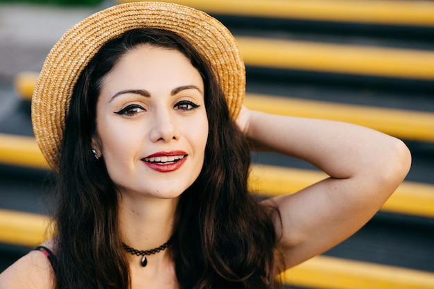 Mujer bonita con ojos brillantes de cabello oscuro y labios pintados de rojo mirando con confianza y con una sonrisa a la cámara con sombrero de paja y collar en el cuello Modelo femenino positivo que tiene vacaciones de verano