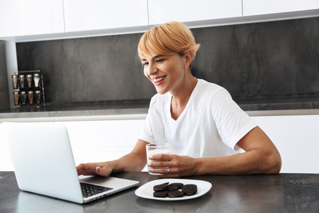 Mujer bonita con ordenador portátil mientras está sentado en la mesa de la cocina, bebiendo leche de un vaso, comiendo galletas