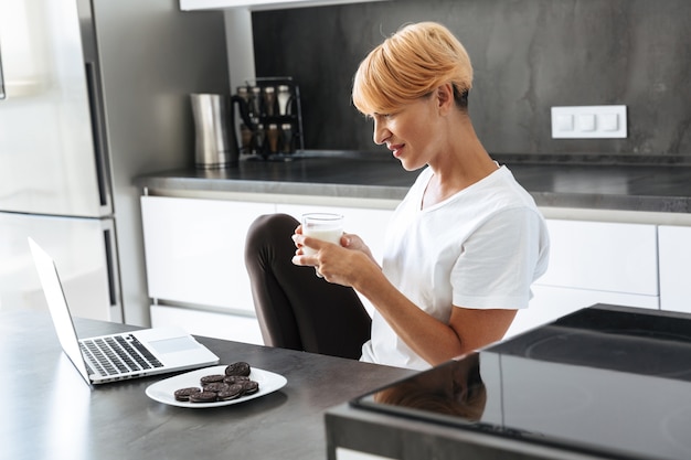 Mujer bonita con ordenador portátil mientras está sentado en la mesa de la cocina, bebiendo leche de un vaso, comiendo galletas