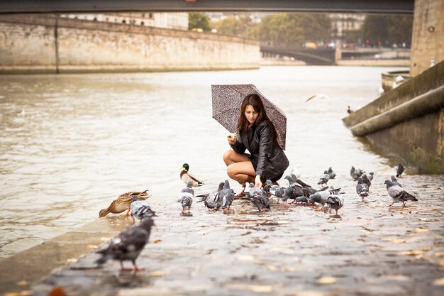 Una mujer bonita con un paraguas alimenta a los pájaros en el terraplén del río.
