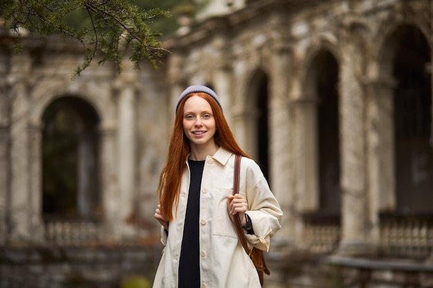 Mujer bonita pelirroja con abrigo en busca de un hermoso lugar histórico, mirando a su alrededor, de pie en contemplación, explorando una nueva área, joven viajero disfruta caminando solo, retrato. viaje, concepto de viaje