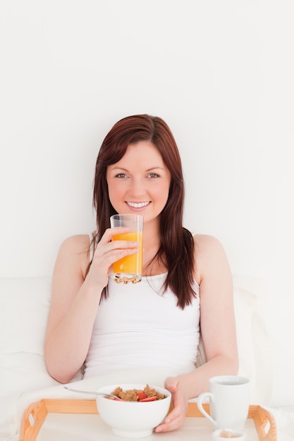 Mujer bonita pelirroja bebiendo un vaso de jugo de naranja mientras está sentado en su cama
