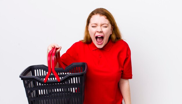 Mujer bonita pelirroja gritando agresivamente mirando muy enojado concepto de cesta de la compra vacía