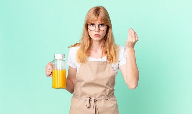 Mujer bonita pelirroja haciendo capice o gesto de dinero, diciéndole que pague con delantal preparando un jugo de naranja