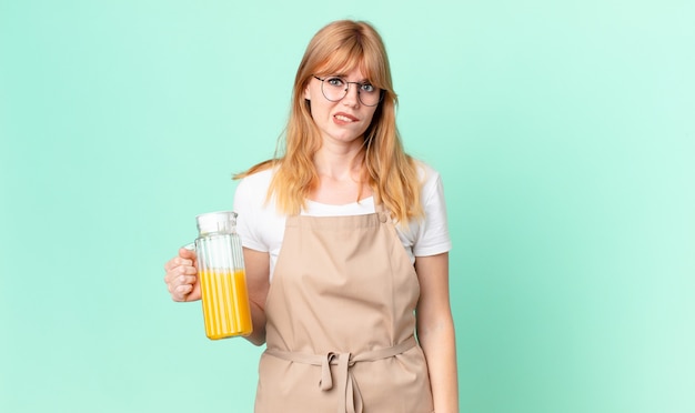 Mujer bonita pelirroja que mira perpleja y confundida con delantal preparando un jugo de naranja