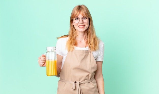 Mujer bonita pelirroja que parece feliz y gratamente sorprendida con delantal preparando un jugo de naranja