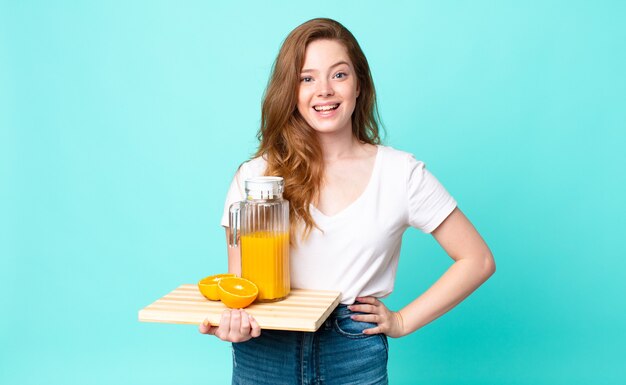 Mujer bonita pelirroja que parece feliz y gratamente sorprendida y sosteniendo un jugo de naranja