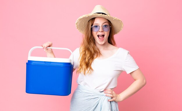 Mujer bonita pelirroja que parece muy conmocionada o sorprendida y sosteniendo un refrigerador portátil de picnic