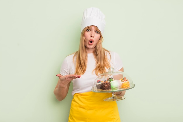 Mujer bonita pelirroja que parece sorprendida y conmocionada con la mandíbula caída sosteniendo un objeto concepto de pasteles caseros