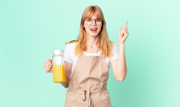 Mujer bonita pelirroja que se siente como un genio feliz y emocionado después de darse cuenta de una idea con un delantal preparando un jugo de naranja