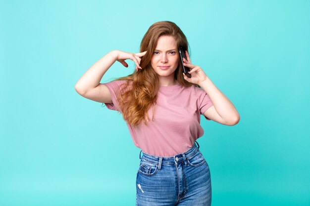 Mujer bonita pelirroja que se siente confundida y perpleja mostrando que estás loco y usando un teléfono inteligente