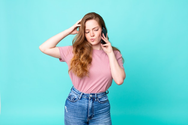 Mujer bonita pelirroja que se siente desconcertada y confundida, rascándose la cabeza y usando un teléfono inteligente