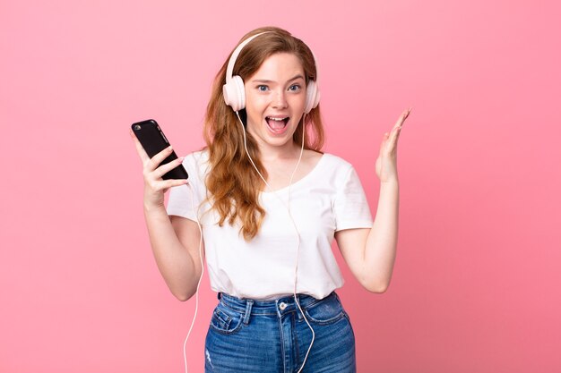 Mujer bonita pelirroja que se siente feliz y asombrada por algo increíble con auriculares y teléfono inteligente