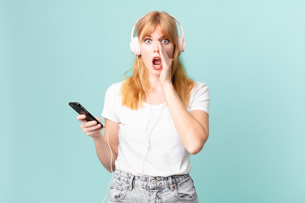 Mujer bonita pelirroja que se siente feliz, dando un gran grito con las manos al lado de la boca y escuchando música con auriculares